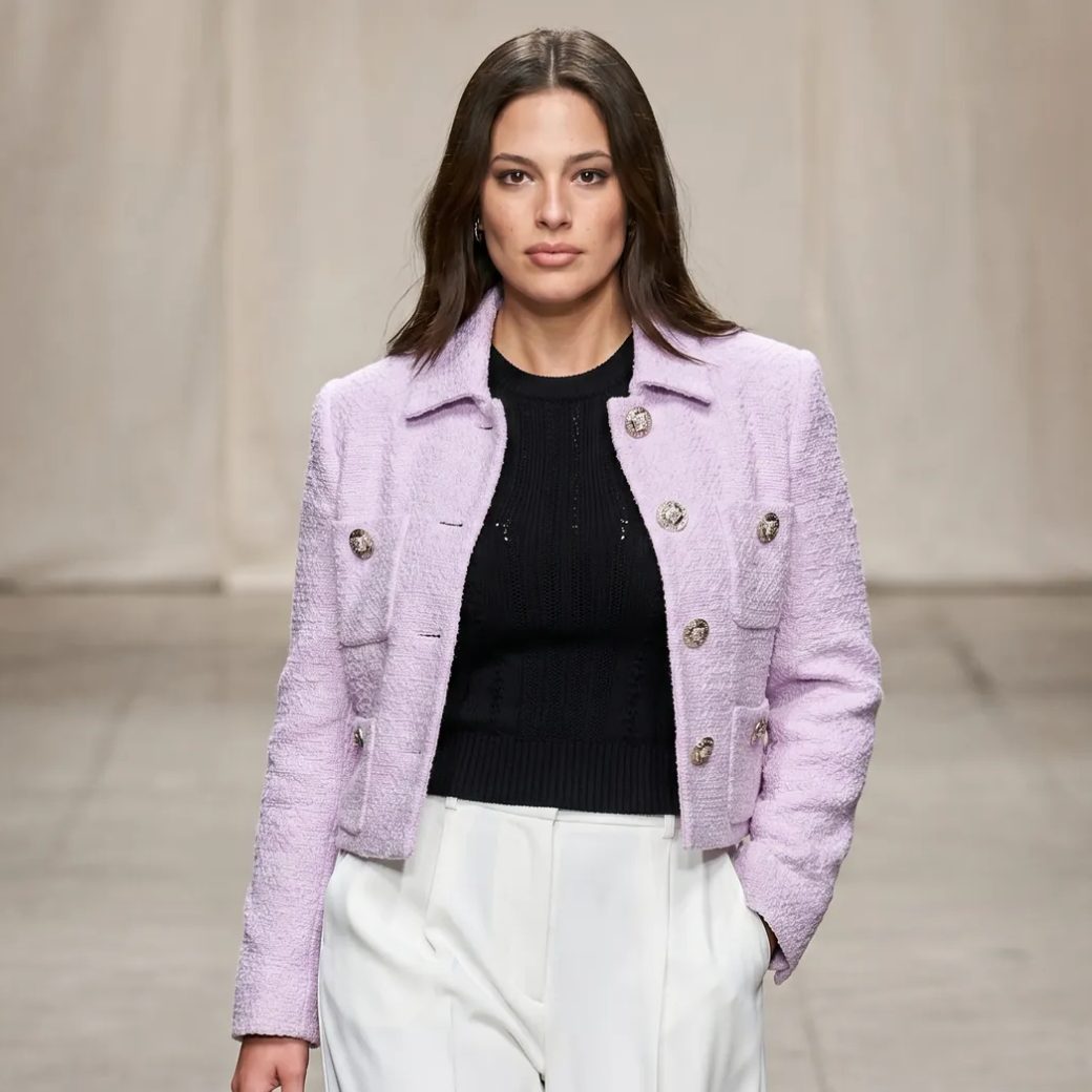 Женщина позирует на подиуме, на ней костюм от Versace: короткий светло-сиреневый жакет с фактурными пуговицами, под ним черный трикотажный топ и широкие белые брюки. Фон нейтральный, освещение мягкое, средний план, уверенный взгляд в камеру.